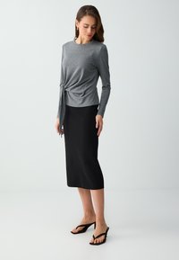 Femme portant un haut gris à manches longues avec un nœud sur le côté, une jupe midi noire et des sandales noires à talons, debout sur un fond blanc uni.
