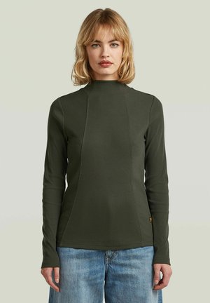 Vrouw met blond haar die een olijfgroene top met lange mouwen en lichtblauwe spijkerbroek draagt, staand tegen een effen lichte achtergrond.