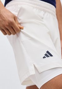 Κοντινό πλάνο ενός ατόμου που τραβά το πλάι ενός λευκού αθλητικού σορτς με ελαστική μέση και μαύρο λογότυπο κοντά στο τελείωμα.