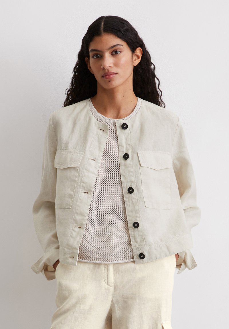 Light beige linen jacket with a round neckline, front buttons, and two chest pockets. Worn over a textured white mesh top.