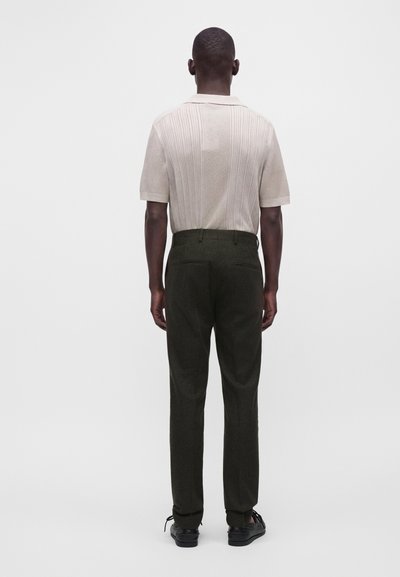 Man standing facing away wearing a short-sleeve textured beige polo shirt and dark tailored pants with black shoes.