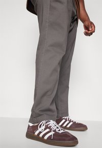 Bordeaux-suède sneakers met witte strepen en bruine zolen, gecombineerd met grijze, losse broek. Sneakers hebben witte veters en getextureerde accenten.