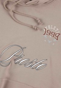 Jasno beżowa bluza z kapturem i sznurkiem, z haftowanym logo "Prosto" w białym kolorze oraz "ATHLET 1999" w czerwonym, z gwiazdkami. Miękka tekstura.