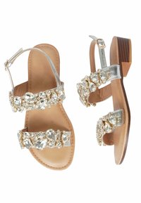 Sandal in argento con due cinturini decorati da strass trasparenti, soletta in pelle beige e tacco in legno marrone. Design con apertura sul retro.