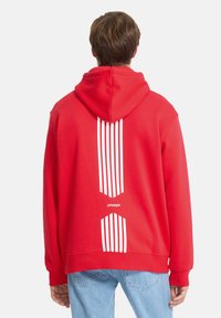 Sweat à capuche rouge avec des rayures verticales blanches à l'arrière, fabriqué en tissu doux. Comprend une capuche ajustable et des poignets côtelés.