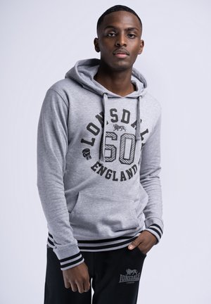 Sudadera gris con gráficos negros, que presenta un bolsillo canguro y cordones. Acentuada con puños y cintura a rayas. Combinada con pantalones negros.