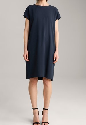 Frau trägt ein knielanges, locker sitzendes dunkelnavyblaues Kleid mit kurzen Ärmeln und schwarzen High Heels mit Knöchelriemen, steht vor einem schlichten Hintergrund.