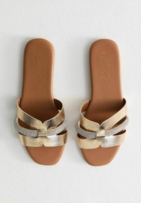 Sandali con cinturini in oro e argento, con una finitura testurizzata, suola piatta marrone e un fiocco decorativo sui cinturini.