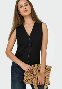 Frau in einer ärmellosen schwarzen, geknöpften Weste und dunklen Jeans, die eine beigen, geflochtenen Handtasche hält, mit goldenen Ringen und Armband als Zubehör.