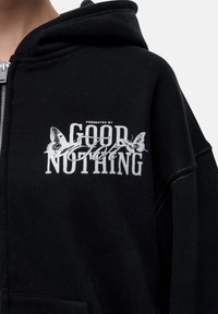 Felpa nera con zip e texture morbida. Presenta una stampa grafica bianca con la scritta "PRESENTED BY GOOD NOTHING" e accenti a forma di farfalla.