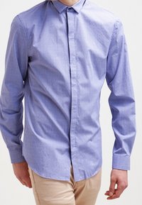 Koszula na guziki z długim rękawem w jasnoniebieskim kolorze o subtelnej fakturze, z kołnierzem i prostym dołem, noszona z beżowymi spodniami.