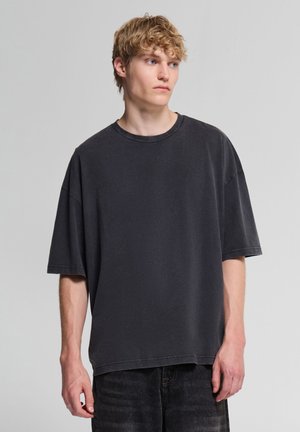 Giovane uomo con capelli biondi e ricci, che indossa una maglietta nera larga e jeans scuri, in piedi davanti a uno sfondo grigio uniforme, guarda verso destra.