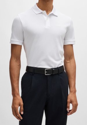 Ein Mann trägt ein eng anliegendes, weißes Kurzarm-Polohemd, das in schwarze, plissierte Hosen gesteckt ist, ergänzt durch einen schwarzen Ledergürtel.