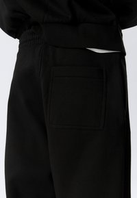 Black sweatpants featuring a smooth fleece texture with an elastic waistband and a rear patch pocket. Simple design with minimal accents.