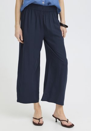 Vrouw draagt marineblauwe broek met wijde pijpen en elastische tailleband, blauw ingestopt shirt, zwarte armband en bruine sandalen met hak.