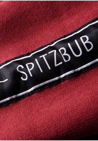 Étiquette en tissu noir avec texte blanc brodé "SPITZBUB" cousue sur un tissu texturé rouge foncé.