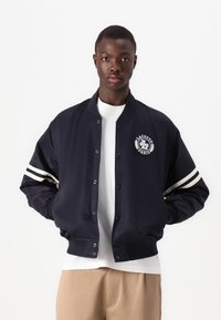 Chaqueta bomber marina con mangas de rayas blancas, cierre de botones a presión y detalle de logo bordado. Tejido suave y ajuste relajado.