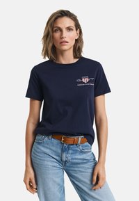 T-shirt en coton bleu marine avec encolure ronde, arborant un logo imprimé, associé à un jean en denim bleu clair et une ceinture marron texturée.