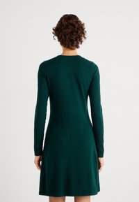 Μακρυμάνικη, σκούρα πράσινη φόρμα με στενή εφαρμογή στο μπούστο και φαρδιά φούστα, που διαθέτει στρογγυλή λαιμόκοψη και λεία υφή.