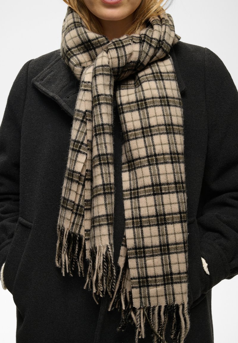 Femme portant une écharpe à carreaux beige et noire avec des franges sur un manteau sombre, mains dans les poches du manteau, sur un fond uni.
