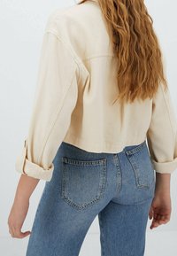 Przycięta beżowa kurtka dżinsowa z podwiniętymi rękawami, posiadająca klasyczny kołnierzyk. Noszona na jasnoniebieskie dżinsy z wysokim stanem z tylnymi kieszeniami.