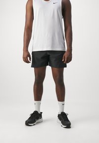 Shorts de sport noirs, t-shirt sans manches blanc, baskets noires avec accents blancs et chaussettes blanches avec logo noir. Design simple et fonctionnel.