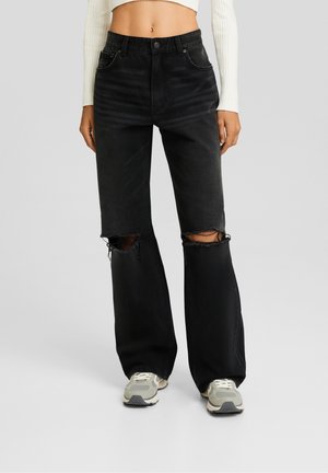 Femme portant un jean noir déchiré à jambes larges, un pull court en maille côtelée blanche et des baskets grises, debout devant un fond uni.
