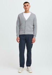 Cardigan en tricot gris avec un col en V et une fermeture à boutons, porté sur un T-shirt blanc, associé à un pantalon sombre et des baskets claires.