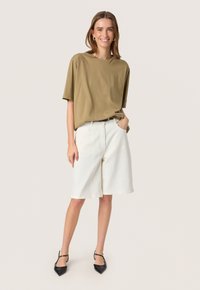 Tricou oversized de culoare verde măslin, asortat cu pantaloni scurți din denim alb cu talie înaltă și pantofi cu vârful ascuțit de culoare neagră, având un design minimalist.
