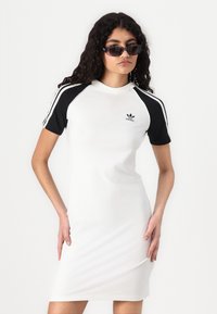White dress with black short sleeves, featuring a round neckline and Adidas logo on the chest. Smooth fabric with sporty design accents.