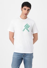 Weißes Baumwoll-T-Shirt mit grünem, schrägem Textlogo "calvin klein" in unterschiedlichen Größen. Kurze Ärmel und Rundhalsausschnitt. Einfaches Design.