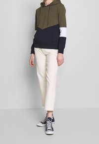 Sudadera con capucha en verde oliva y azul marino, con un diseño de flecha en blanco. Combinada con pantalones en un tono blanco roto y zapatillas azul marino.