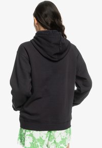 Sweat à capuche noir avec une capuche spacieuse, une texture douce et une coupe ample. Présente des poignets et un ourlet côtelés, vu de dos avec une jupe verte à motifs.