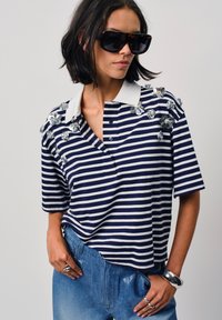 Marineblauwe en witte gestreepte polo met korte mouwen, een witte kraag en versierde bloemen op de schouder. Draagt met lichte spijkerbroeken.