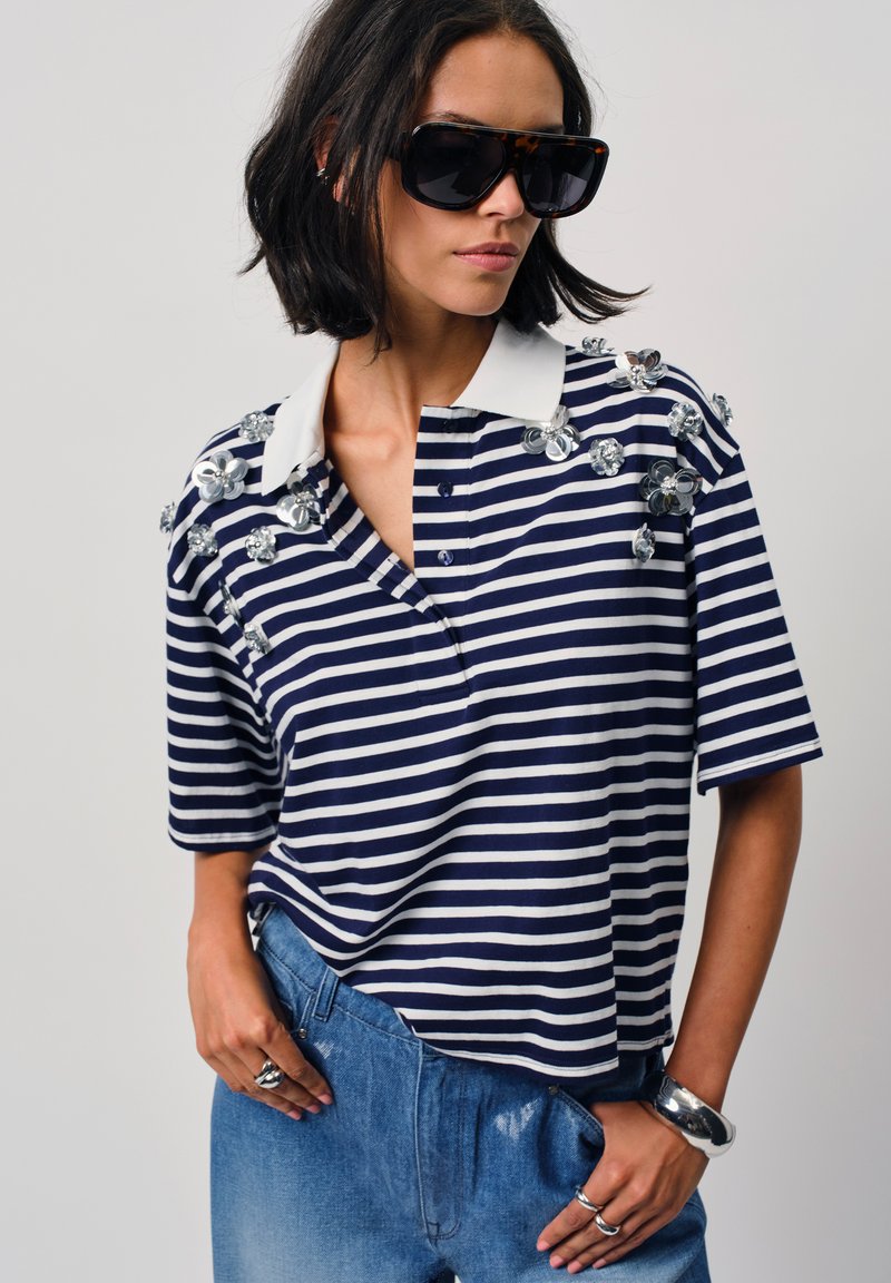 Marineblauwe en witte gestreepte polo met korte mouwen, een witte kraag en versierde bloemen op de schouder. Draagt met lichte spijkerbroeken.