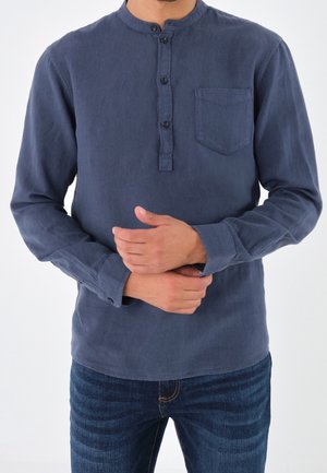 Uomo che indossa una maglia henley blu navy a maniche lunghe con taschino sul petto e jeans blu, mani giunte davanti all’altezza della vita.