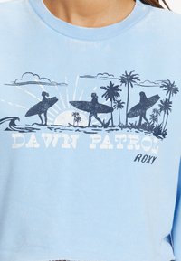 Felpa crop azzurra chiara con una grafica retro di surf che presenta palme, sole e il testo "DAWN PATROL" in bianco e blu navy. Materiale in cotone.
