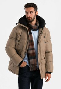 Chaqueta de plumas beige con capucha negra, que presenta botones a presión, bolsillos laterales y una superficie texturizada; combinada con una bufanda de cuadros y un suéter azul.
