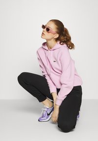 Lys rosa hettegenser med snor og grønn logo, sammen med sorte joggebukser og lilla/hvite joggesko. Modellen har på seg rosa solbriller.