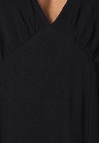 Robe noire présentant un décolleté en V, un détail froncé aux épaules et un tissu texturé avec des motifs subtils.