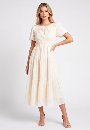 Femme debout dans une robe midi crème à volants avec des manches bouffantes et des talons à bride à la cheville, sur un fond uni.