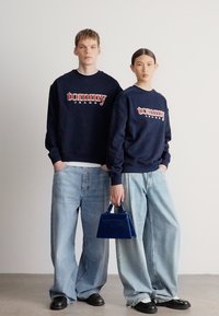 Námornícke mikiny s červeným nápisom „Tommy Jeans“, široké svetlomodré džínsy a modrá kabelka. Tkanina pôsobí mäkko a má hladkú textúru.