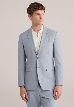 Jonge man die een lichtblauw colbert en bijpassende broek draagt over een wit shirt met rits, kijkend naar de zijkant.