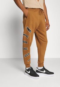 Man som bär bruna träningsoverallsbyxor med texten "JORDAN" nerför vänster ben och svart-vita Nike-sneakers, stående mot en vit bakgrund.