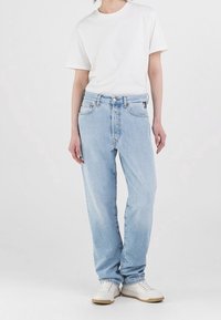 Person som bär en enkel vit t-shirt, ljusblå högmidjade jeans med knäppning framtill och vita sneakers, stående mot en neutral bakgrund.