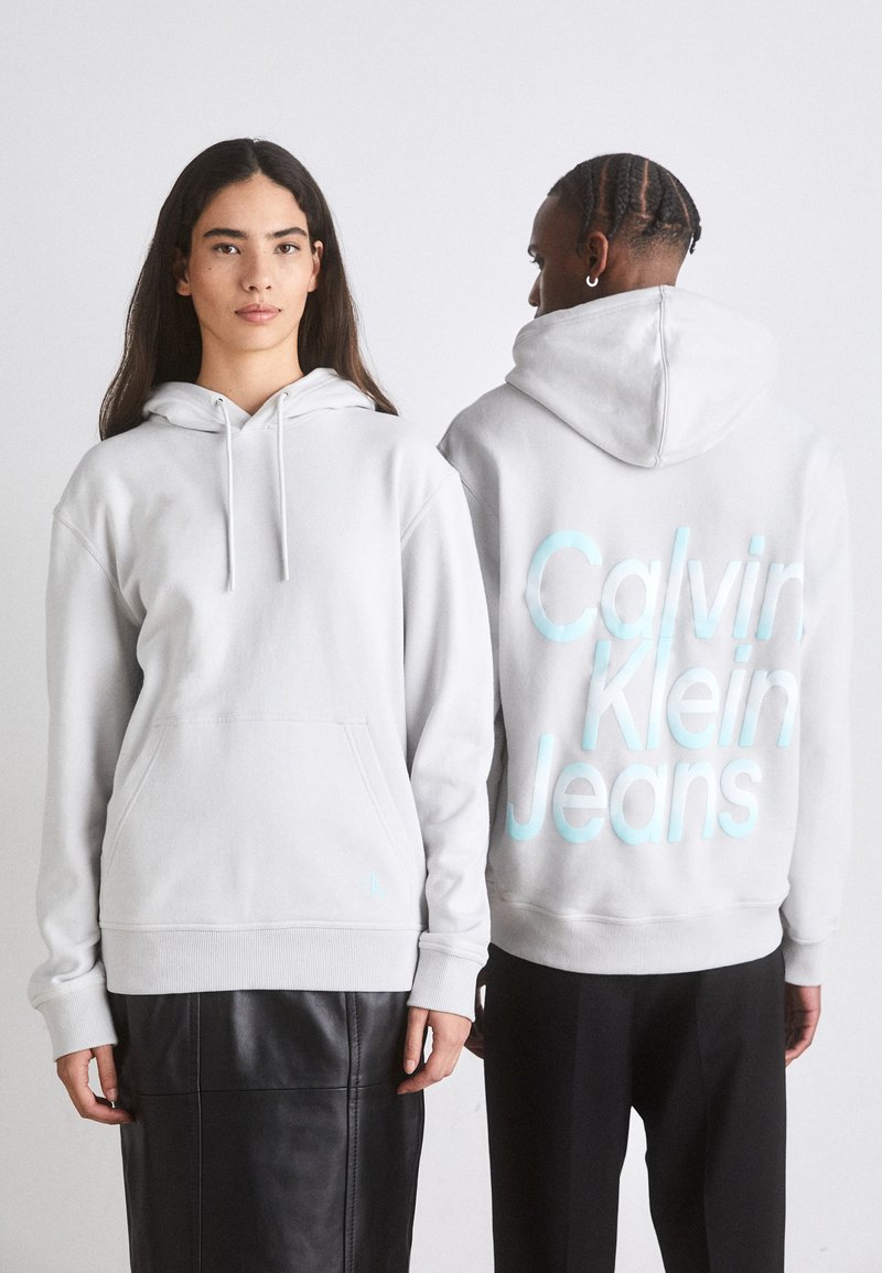 Hoodie grigio chiaro con coulisse, tasca frontale e dettaglio del logo. Il retro presenta una grande stampa aqua "Calvin Klein Jeans". Texture morbida in felpa.