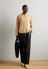 Cămașă bej cu nasturi, dotată cu două buzunare frontale, asortată cu pantaloni largi din piele neagră. Modelul ține în mână o geantă de umăr neagră și poartă tocuri ușoare.