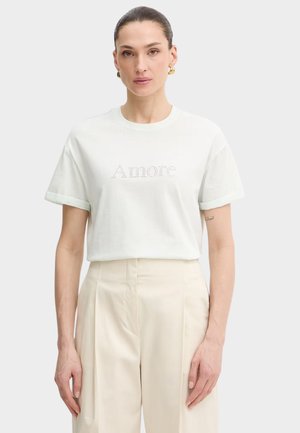 Donna che indossa una t-shirt bianca con la scritta "Amore" in piccoli fori e pantaloni crema a vita alta, in piedi davanti a uno sfondo bianco.