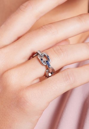 Zilverkleurige ring met onderling verbonden ketting schakels in verschillende vormen, gepolijste textuur en een gladde afwerking, gedragen aan een vinger.