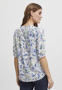 Blouse florale avec un col, des manches bouffantes et une coupe décontractée. Les couleurs incluent le bleu, le violet et le vert sur un fond clair.